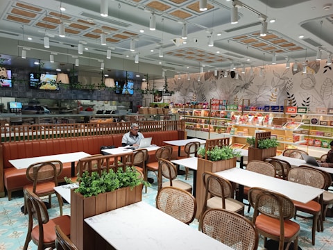 A modern and well-lit cafe with neatly arranged tables and chairs. The walls are decorated with botanical designs, and there is a variety of packaged products on shelves along the wall. The seating area features brown leather benches and wooden chairs. Green plants are used as decor on the tables. There are a couple of people seated, one of whom is working on a laptop.