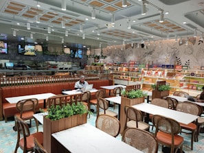 A modern and well-lit cafe with neatly arranged tables and chairs. The walls are decorated with botanical designs, and there is a variety of packaged products on shelves along the wall. The seating area features brown leather benches and wooden chairs. Green plants are used as decor on the tables. There are a couple of people seated, one of whom is working on a laptop.