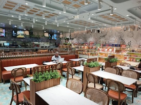 A modern and well-lit cafe with neatly arranged tables and chairs. The walls are decorated with botanical designs, and there is a variety of packaged products on shelves along the wall. The seating area features brown leather benches and wooden chairs. Green plants are used as decor on the tables. There are a couple of people seated, one of whom is working on a laptop.
