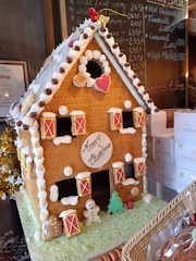 A gingerbread house intricately decorated with icing, candies, and cookies. The roof is adorned with chocolate buttons and white icing resembling snow. A heart-shaped candy is placed near the roof's peak. Windows are framed with yellow and red icing, and 'Merry Christmas' is inscribed on the front in chocolate. Various small decorative figures, including gingerbread men and a Christmas tree, are placed around the house.