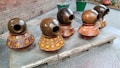 A group of five decorative clay pots with glossy brown tops and colorful bases featuring intricate patterns and designs. The pots are lined up on a concrete slab with a patterned walkway and brick wall in the background.