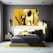 A modern bedroom with a large, abstract painting in yellow, white, and black shades hanging above the bed. The decor includes matching yellow accents in the pillows, lamp shades, and a rug. The room features a contemporary style with a gray wall, wooden nightstand, and a tall green plant.