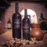 Two wine bottles labeled 'Rosselli Reserve' stand on a wooden table surrounded by several corks. A copper jug is placed next to the bottles, with a blurred background of a rustic wine cellar featuring stone walls and additional wine bottles.