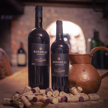 Two wine bottles labeled 'Rosselli Reserve' stand on a wooden table surrounded by several corks. A copper jug is placed next to the bottles, with a blurred background of a rustic wine cellar featuring stone walls and additional wine bottles.