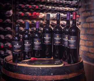 A collection of wine bottles is arranged on a wooden barrel, set against a backdrop of a wine cellar with shelves of bottles. The bottles are labeled 'Rosselli Reserve' and contain different varieties of wine. A pair of red-handled tongs and a server are placed in front of the bottles.