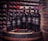 A collection of wine bottles is arranged on a wooden barrel, set against a backdrop of a wine cellar with shelves of bottles. The bottles are labeled 'Rosselli Reserve' and contain different varieties of wine. A pair of red-handled tongs and a server are placed in front of the bottles.
