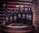 A collection of wine bottles is arranged on a wooden barrel, set against a backdrop of a wine cellar with shelves of bottles. The bottles are labeled 'Rosselli Reserve' and contain different varieties of wine. A pair of red-handled tongs and a server are placed in front of the bottles.