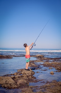 a young boy standing on top of a rocky beach holding a fishing pole