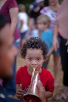 A young child with curly hair is playing a brass or copper-colored instrument, possibly a trumpet or horn. They are wearing a red shirt and surrounded by a blurred background where other people are present, creating a sense of a crowded outdoor gathering or event.