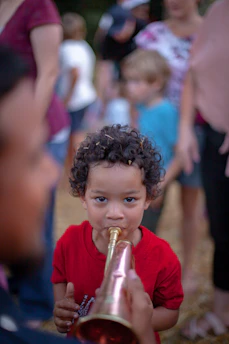 A young child with curly hair is playing a brass or copper-colored instrument, possibly a trumpet or horn. They are wearing a red shirt and surrounded by a blurred background where other people are present, creating a sense of a crowded outdoor gathering or event.