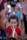 A young child with curly hair is playing a brass or copper-colored instrument, possibly a trumpet or horn. They are wearing a red shirt and surrounded by a blurred background where other people are present, creating a sense of a crowded outdoor gathering or event.