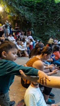 A young child in a green shirt reaches out and holds onto an adult's arm in an outdoor setting filled with people sitting and standing. The background has lush green foliage and a warmly lit atmosphere. There are multiple people of various ages engaging in different activities and conversations.
