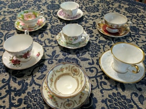 An arrangement of six ornate teacups with matching saucers is displayed on an intricately patterned dark blue lace tablecloth. Each teacup features a unique floral design with vibrant colors and gold accents.