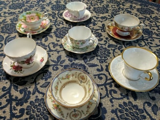 An arrangement of six ornate teacups with matching saucers is displayed on an intricately patterned dark blue lace tablecloth. Each teacup features a unique floral design with vibrant colors and gold accents.
