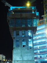A construction site at night featuring a partially constructed building with visible concrete and metal framework. The scene is illuminated by artificial lighting, with a crane visible in the background. The structure is surrounded by scaffolding and there are layers of floors under construction.