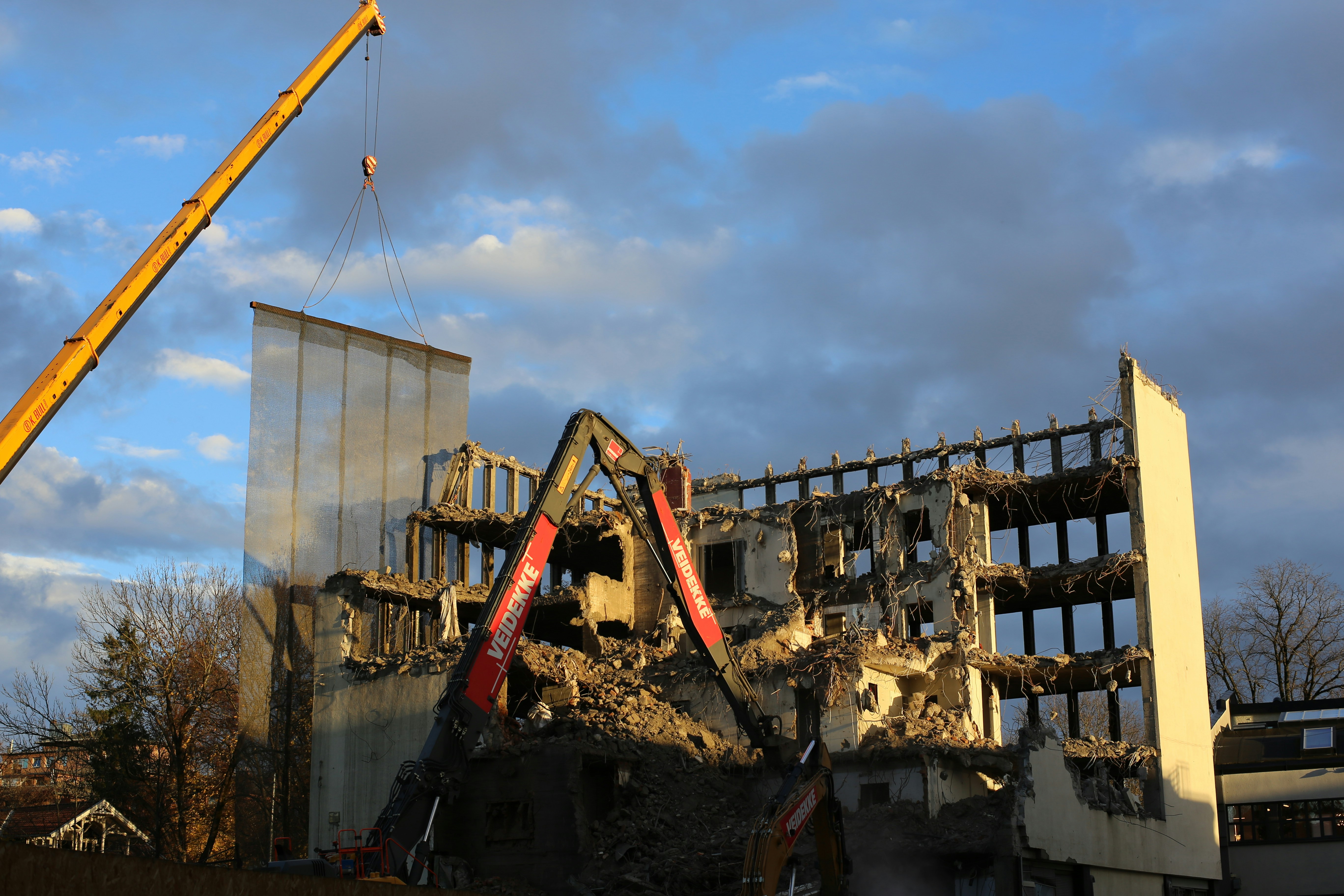A partially demolished building with several floors exposed and crumbling rubble around. Two large construction cranes are positioned next to the building, assisting in the demolition process. The sky is partially cloudy, with sections of clear blue visible.