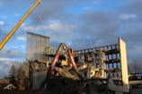 A partially demolished building with several floors exposed and crumbling rubble around. Two large construction cranes are positioned next to the building, assisting in the demolition process. The sky is partially cloudy, with sections of clear blue visible.