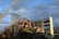 A partially demolished building with several floors exposed and crumbling rubble around. Two large construction cranes are positioned next to the building, assisting in the demolition process. The sky is partially cloudy, with sections of clear blue visible.