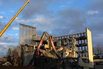 A partially demolished building with several floors exposed and crumbling rubble around. Two large construction cranes are positioned next to the building, assisting in the demolition process. The sky is partially cloudy, with sections of clear blue visible.