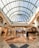 A spacious and elegant shopping mall interior featuring a high, glass-domed ceiling that allows natural light to illuminate the area. The floor is made of polished marble with various patterns. People are leisurely walking and shopping, with several store fronts and a café visible in the background.