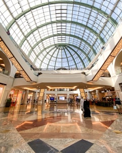 A spacious and elegant shopping mall interior featuring a high, glass-domed ceiling that allows natural light to illuminate the area. The floor is made of polished marble with various patterns. People are leisurely walking and shopping, with several store fronts and a café visible in the background.