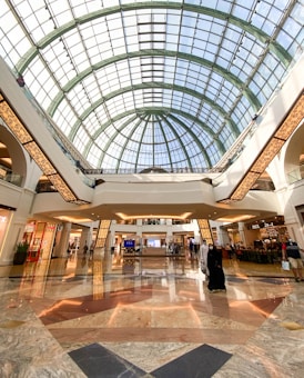 A spacious and elegant shopping mall interior featuring a high, glass-domed ceiling that allows natural light to illuminate the area. The floor is made of polished marble with various patterns. People are leisurely walking and shopping, with several store fronts and a caf&eacute; visible in the background.