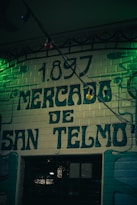A vintage-style sign with the year 1897 written above the words Mercado de San Telmo, painted on a brick wall with a dark and moody ambiance. The image is slightly dim with green lighting adding to the nostalgic and historical feeling.