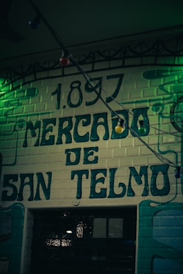 A vintage-style sign with the year 1897 written above the words Mercado de San Telmo, painted on a brick wall with a dark and moody ambiance. The image is slightly dim with green lighting adding to the nostalgic and historical feeling.