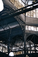 An intricate metal framework supports a large industrial ceiling. The structure includes various beams and ornate designs with light filtering through slotted windows. The scene is reminiscent of an old factory or market hall with its heavy reliance on metal construction.