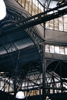 An intricate metal framework supports a large industrial ceiling. The structure includes various beams and ornate designs with light filtering through slotted windows. The scene is reminiscent of an old factory or market hall with its heavy reliance on metal construction.