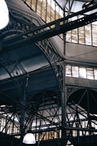 An intricate metal framework supports a large industrial ceiling. The structure includes various beams and ornate designs with light filtering through slotted windows. The scene is reminiscent of an old factory or market hall with its heavy reliance on metal construction.
