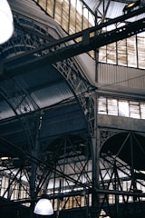 An intricate metal framework supports a large industrial ceiling. The structure includes various beams and ornate designs with light filtering through slotted windows. The scene is reminiscent of an old factory or market hall with its heavy reliance on metal construction.