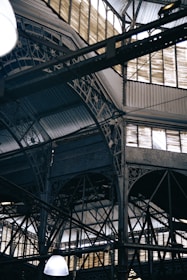 An intricate metal framework supports a large industrial ceiling. The structure includes various beams and ornate designs with light filtering through slotted windows. The scene is reminiscent of an old factory or market hall with its heavy reliance on metal construction.