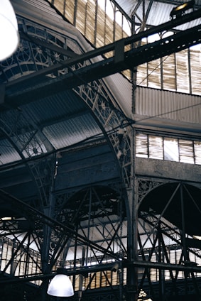 An intricate metal framework supports a large industrial ceiling. The structure includes various beams and ornate designs with light filtering through slotted windows. The scene is reminiscent of an old factory or market hall with its heavy reliance on metal construction.