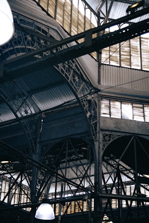 An intricate metal framework supports a large industrial ceiling. The structure includes various beams and ornate designs with light filtering through slotted windows. The scene is reminiscent of an old factory or market hall with its heavy reliance on metal construction.