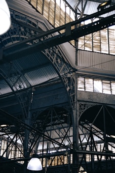 An intricate metal framework supports a large industrial ceiling. The structure includes various beams and ornate designs with light filtering through slotted windows. The scene is reminiscent of an old factory or market hall with its heavy reliance on metal construction.
