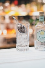 A tall glass filled with ice cubes, accompanied by a perforated spoon resting on top. Next to the glass is a bottle of gin with a decorative label. The background is softly blurred, featuring warm, ambient lighting suggesting a bar setting.