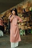 A woman is standing and smiling in front of a display of woven handbags and hats. She is wearing a pink tunic with white pants and a black hat. The background features numerous bags hung on a structure and several hats stacked on stools, indicative of a market or shop setting.