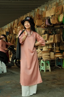 A woman is standing and smiling in front of a display of woven handbags and hats. She is wearing a pink tunic with white pants and a black hat. The background features numerous bags hung on a structure and several hats stacked on stools, indicative of a market or shop setting.