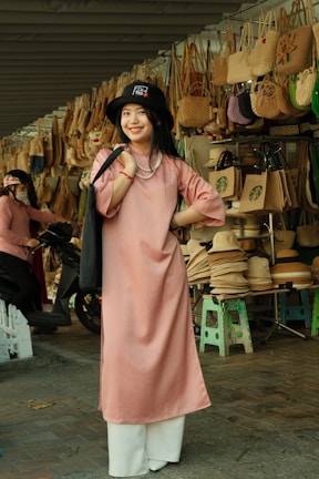 A woman is standing and smiling in front of a display of woven handbags and hats. She is wearing a pink tunic with white pants and a black hat. The background features numerous bags hung on a structure and several hats stacked on stools, indicative of a market or shop setting.