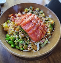 A bowl filled with fresh ingredients, including slices of raw salmon arranged on top. The dish contains edamame, avocado, diced mango, and other colorful vegetables, all drizzled with a savory sauce and sprinkled with sesame seeds.
