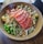 A bowl filled with fresh ingredients, including slices of raw salmon arranged on top. The dish contains edamame, avocado, diced mango, and other colorful vegetables, all drizzled with a savory sauce and sprinkled with sesame seeds.