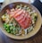 A bowl filled with fresh ingredients, including slices of raw salmon arranged on top. The dish contains edamame, avocado, diced mango, and other colorful vegetables, all drizzled with a savory sauce and sprinkled with sesame seeds.
