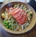 A bowl filled with fresh ingredients, including slices of raw salmon arranged on top. The dish contains edamame, avocado, diced mango, and other colorful vegetables, all drizzled with a savory sauce and sprinkled with sesame seeds.