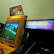 A vintage arcade setup featuring two arcade machines. The first is a yellow Crazy Taxi arcade machine with a steering wheel, featuring vibrant signage at the top displaying 'Crazy Taxi High Roller' with flames. The screen shows a driving game landscape with roads and trees. Next to it is another machine with a sign that reads 'Electronic Experience' with a colorful design, though its screen is not lit.