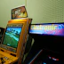 A vintage arcade setup featuring two arcade machines. The first is a yellow Crazy Taxi arcade machine with a steering wheel, featuring vibrant signage at the top displaying 'Crazy Taxi High Roller' with flames. The screen shows a driving game landscape with roads and trees. Next to it is another machine with a sign that reads 'Electronic Experience' with a colorful design, though its screen is not lit.