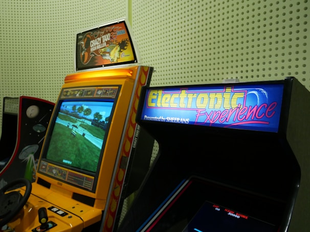 A vintage arcade setup featuring two arcade machines. The first is a yellow Crazy Taxi arcade machine with a steering wheel, featuring vibrant signage at the top displaying 'Crazy Taxi High Roller' with flames. The screen shows a driving game landscape with roads and trees. Next to it is another machine with a sign that reads 'Electronic Experience' with a colorful design, though its screen is not lit.