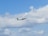 An airplane with branding and logo visible on its side flies amidst a partly cloudy sky, with a few birds soaring nearby. The scene suggests an in-flight journey of the aircraft against a backdrop of fluffy clouds.
