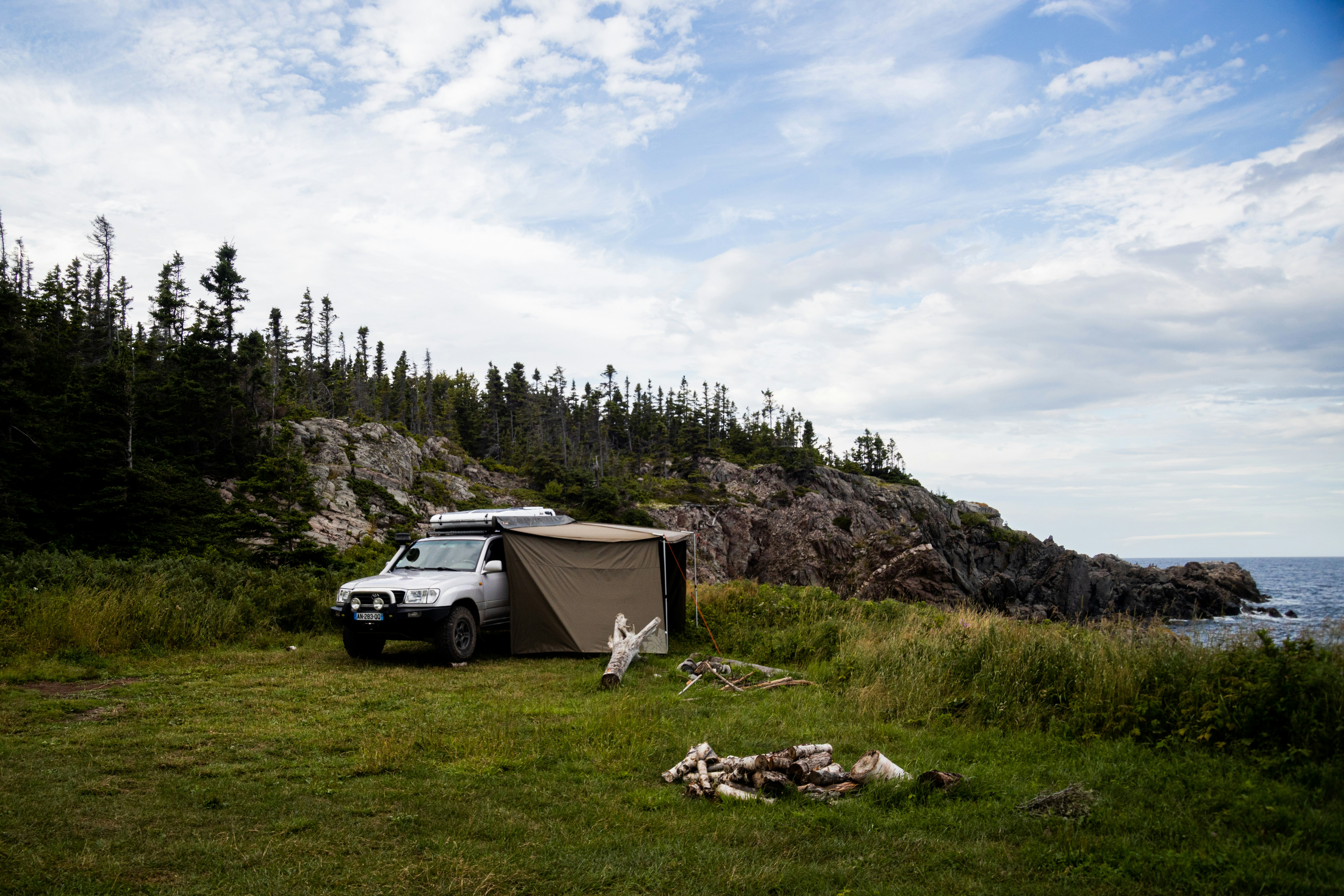 Silver SUV parked on a grassy headland above a rocky shore, a side awning with fabric walls attached along its passenger side and spruce trees behind