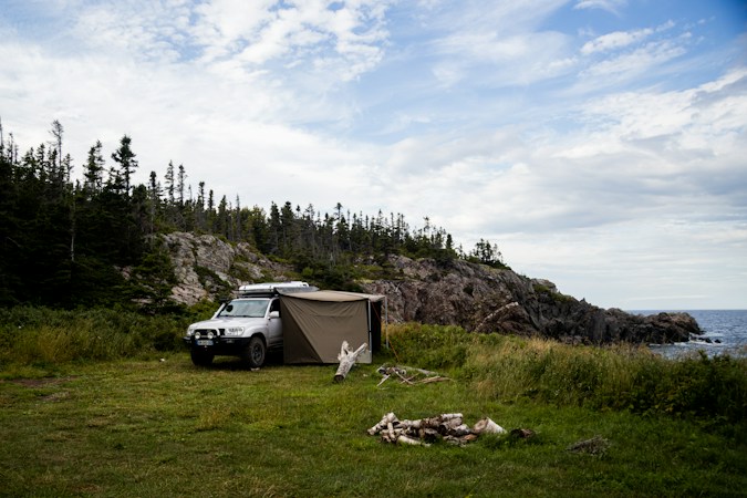 Silver SUV parked on a grassy headland above a rocky shore, a side awning with fabric walls attached along its passenger side and spruce trees behind